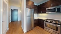 This contemporary high-rise residence is designed with clean lines and modern finishes throughout. The entry hallway, finished with warm wood-look flooring, leads into the main living areas and is flanked by a louvered closet door offering generous storage. The kitchen is a standout feature, outfitted with rich dark wood cabinetry, light quartz countertops, a classic subway tile backsplash, and a full suite of stainless steel appliances including a gas range, over-the-range microwave, and full-size refrigerator. The open layout connects seamlessly to the living area, which enjoys expansive floor-to-ceiling windows framing impressive panoramic city views. Both bedrooms are well-proportioned — one featuring hardwood-style flooring and large windows with sweeping cityscape vistas, the other carpeted and served by a dedicated closet. Two full bathrooms maintain the home's modern aesthetic, each featuring a tub-shower combination with tile surrounds, wood-toned vanity cabinetry, and clean white countertops. The overall ambiance is bright, airy, and contemporary, with abundant natural light flooding the space through oversized windows that showcase the urban skyline beyond.