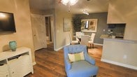 This single-floor residence offers an open, connected layout where the living and dining areas flow naturally into one another. Rich hardwood flooring runs throughout, providing warmth and continuity across the space. The living area features a tufted blue accent chair paired with a decorative throw pillow, anchored beneath a ceiling fan that adds both function and a casual charm to the room.

The dining area is defined by a glass-top table surrounded by upholstered chairs, accented by a decorative wall mirror and a tall indoor plant that adds a natural, organic touch. A track lighting fixture overhead provides focused illumination, complementing the ambient light from a nearby window. The walls are finished in a two-tone palette of warm beige and cool gray, creating a subtle yet visually interesting contrast throughout the space.

Adjacent to the dining area, a partial view of what appears to be a kitchen counter with granite-style surfaces hints at additional functional space nearby. A hallway with louvered bi-fold doors leads toward a bathroom, suggesting well-organized storage and private spaces just off the main living area. Overall, the space presents a comfortable, well-appointed interior with thoughtful design details.