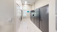 Located at 13625 Haven Ridge Ln in Charlotte, NC, this single-story package and mail center is designed to provide residents with a secure and convenient parcel management experience. The facility is anchored by a bank of Luxer One smart package lockers lining the walls, offering multiple compartment sizes to accommodate deliveries of varying dimensions. The space is monitored around the clock with 24/7 video surveillance, as indicated by posted signage, ensuring the safety and security of all parcels. Light-toned porcelain tile flooring runs throughout, complementing the neutral warm-white walls and crisp white baseboard trim for a clean, modern aesthetic. Natural light filters in through windows fitted with white horizontal blinds, brightening the interior and creating a welcoming atmosphere. A dedicated counter area features rich navy-blue shaker-style cabinetry with gold hardware and a white quartz countertop, providing a functional workspace for managing packages and mail. The hallway connects to adjacent rooms, one of which displays abstract wall art, adding a touch of character to the utilitarian space. Additional utility elements, including a stainless steel waste receptacle and a gray utility cabinet, are thoughtfully integrated without disrupting the overall clean design. The facility is well-ventilated with ceiling vents and recessed lighting, ensuring comfort during use.