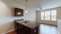 Located at 1200 E Parmer Ln in Austin, TX, this single-floor residential unit offers a practical and cohesive layout designed for comfortable everyday living. The open-concept kitchen is a central highlight, featuring dark espresso cabinetry with granite-style countertops, a peninsula with an undermount sink, pendant lighting, and black appliances including a range and microwave. The peninsula provides a natural transition into the adjacent dining and living areas, which are unified by warm wood-look flooring that extends throughout the unit. Ceiling fans are present in multiple rooms, supporting airflow and comfort. The living area is bright and open, benefiting from large windows fitted with horizontal blinds that allow ample natural light while maintaining privacy. A generously sized bedroom continues the warm flooring aesthetic and features a wide bank of windows with outdoor views, a ceiling fan, and plenty of wall space for flexible furniture arrangements. The bathroom is well-appointed with a dark wood vanity, granite-style countertop, a large mirror with overhead lighting, and matching flooring. The overall color palette of neutral tones and warm wood finishes creates a cohesive and inviting atmosphere throughout the unit.
