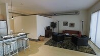 This single-floor space showcases a contemporary, open-concept layout with a distinctly urban, industrial character. Exposed ductwork runs along the ceiling, adding an authentic loft-style aesthetic that complements the clean white walls and light hardwood flooring found throughout. The living area is anchored by a set of sleek black lounge chairs and a rich leather sofa arranged around a dark coffee table, all resting atop a patterned area rug that adds texture and warmth to the space. Framed artwork adorns the walls, contributing a curated, gallery-like quality to the room. A dark wood media console serves as a functional focal point, accompanied by a tall potted plant that introduces a natural element and visual contrast. A woven basket adds a subtle organic touch nearby. The dining area features a high-top table paired with modern metal barstools, offering a casual yet stylish spot for meals or conversation. The open flow between the dining and living zones creates a sense of spaciousness, while the cohesive use of dark furnishings against light walls and floors strikes a balanced, sophisticated tone throughout the space.