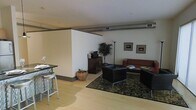 This single-floor space showcases a contemporary, open-concept layout with a distinctly urban, industrial character. Exposed ductwork runs along the ceiling, adding an authentic loft-style aesthetic that complements the clean white walls and light hardwood flooring found throughout. The living area is anchored by a set of sleek black lounge chairs and a rich leather sofa arranged around a dark coffee table, all resting atop a patterned area rug that adds texture and warmth to the space. Framed artwork adorns the walls, contributing a curated, gallery-like quality to the room. A dark wood media console serves as a functional focal point, accompanied by a tall potted plant that introduces a natural element and visual contrast. A woven basket adds a subtle organic touch nearby. The dining area features a high-top table paired with modern metal barstools, offering a casual yet stylish spot for meals or conversation. The open flow between the dining and living zones creates a sense of spaciousness, while the cohesive use of dark furnishings against light walls and floors strikes a balanced, sophisticated tone throughout the space.