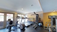 This fitness center offers a dedicated space for residents to pursue their health and wellness goals. The room is thoughtfully designed to maximize functionality, housing a selection of cardio equipment including treadmills and an elliptical trainer, alongside a multi-station cable machine suitable for a range of strength-training exercises. A stability ball rounds out the available equipment, supporting core and flexibility training. The space is brightened by natural light streaming in through large windows and a glass door, which also provide views of the outdoor surroundings and create an open, airy atmosphere. Recessed lighting in the smooth ceiling ensures the room remains well-lit at all times. A dynamic wall mural depicting silhouetted runners against a warm sunset backdrop adds a bold, motivating visual element and energizes the overall ambiance. The dark flooring provides a durable, sport-appropriate surface underfoot. The layout is clean and organized, allowing for comfortable movement between equipment. An easily accessible exit door enhances safety and convenience. Overall, the fitness center delivers a practical and inspiring environment for a complete workout experience.
