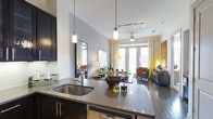 This 1-bedroom, 1-bathroom residence is designed with a modern, open-concept layout that maximizes both space and functionality. The kitchen serves as the heart of the home, featuring dark cabinetry, sleek stone countertops, a stainless steel sink, a dishwasher, and a peninsula that flows seamlessly into the dining and living areas. Pendant lighting and a track lighting system overhead add both task illumination and ambient warmth to the space. The living area is bright and inviting, benefiting from large floor-to-ceiling windows and French doors that flood the room with natural light and offer access to the outdoors. A ceiling fan contributes to the comfortable atmosphere. Rich, dark hardwood-style flooring runs throughout the main living space, grounding the contemporary aesthetic and providing a warm contrast to the light-toned walls. The overall design palette — combining dark wood tones, neutral walls, and modern fixtures — creates a sophisticated yet welcoming ambiance. The open sightlines between the kitchen, dining, and living areas enhance the sense of spaciousness, making this single-floor residence feel open, airy, and well-connected throughout.