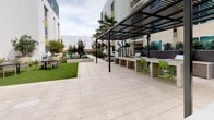 This outdoor courtyard is a thoughtfully designed communal space nestled within the grounds of a modern multi-story residential building. The courtyard is organized into several distinct zones, each offering a unique setting for relaxation and social gathering. A lush artificial turf lawn serves as a central green anchor, surrounded by dining tables and chairs that invite casual outdoor meals. Mature trees and a variety of tropical plantings, including sago palms and ornamental shrubs, are arranged in large planters and landscaped beds, providing greenery and a sense of natural enclosure throughout the space. A standout feature is the sleek metal pergola structure, which shelters a built-in outdoor kitchen and bar counter equipped with a grill and bar-height seating with bright green chairs — an ideal setup for outdoor entertaining. Across the courtyard, vibrant turquoise Adirondack chairs are arranged around a casual lounge area, adding a pop of color. A covered walkway with tile flooring and overhead lighting connects areas of the courtyard and provides a sheltered passage lined with potted tropical plants. The surrounding building facades, finished in neutral tones with teal and yellow accents, frame the courtyard and contribute to its contemporary aesthetic.