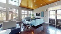 This well-appointed single-floor communal space is designed to impress with its open layout and thoughtful design details throughout. The main lounge area commands attention with soaring double-height ceilings clad in warm wood paneling, which contrast beautifully against the high-gloss, deep-toned polished concrete floors below. Floor-to-ceiling windows line the walls, flooding the interior with abundant natural light while offering views of the surrounding landscaped grounds and outdoor seating areas.

The lounge is furnished with a mix of contemporary seating, including patterned teal sofas, sleek upholstered benches, and accent chairs arranged around a glass coffee table, creating a welcoming gathering atmosphere. A wall-mounted flat-screen television anchors one side of the room, complemented by tasteful artwork in coordinating teal tones.

Adjacent to the lounge, a fully equipped kitchen features stainless steel appliances, dark granite countertops, mosaic tile detailing, and a bar-height counter with seating for multiple guests. Pendant lighting illuminates the space elegantly. A pool table visible in the aerial view further enhances the entertainment-focused programming of this versatile space. Small café-style tables positioned near the windows offer intimate seating with serene garden views.