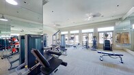 This single-floor fitness center offers a comprehensive and thoughtfully designed workout facility. The space is divided into distinct functional zones, providing ample room for a full range of exercise activities. The cardio area is lined with treadmills, elliptical trainers, and stair climbers, all positioned to take advantage of the abundant natural light streaming through a row of large windows and glass-paneled doors along the exterior wall. Ceiling fans overhead contribute to a comfortable workout environment.

The strength training section features a multi-station cable machine, a selection of weight benches, and a well-stocked dumbbell rack with a wide range of free weights mounted on red wall brackets. Floor-to-ceiling mirrors span entire walls throughout the space, visually expanding the room and allowing users to monitor their form during exercise.

The entry area makes a refined first impression with its soaring ceilings, elegant marble tile flooring, warm textured wall coverings, decorative wainscoting, and a built-in water and bottle-filling station set against a mosaic tile accent wall. Pendant and recessed lighting throughout the facility maintain a bright, welcoming atmosphere. The flooring within the workout areas consists of durable, light-toned carpet tiles, providing both comfort and traction.