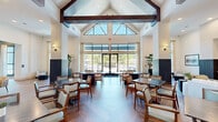 This expansive single-floor community clubhouse offers a generous footprint thoughtfully designed for gathering, dining, and relaxation. The architectural centerpiece is a dramatic vaulted ceiling adorned with bold, dark-stained wood beams that rise to impressive heights, paired with large clerestory windows that flood the interior with natural light and create an airy, open atmosphere.

The dining area features a herringbone-patterned hardwood floor and is lined with neatly arranged tables and cushioned chairs, all bathed in natural light streaming through floor-to-ceiling windows and glass doors that open to an outdoor courtyard. The living area offers comfortable lounge seating and connects seamlessly to a stylish bar nook, highlighted by a striking marble-topped island with a navy blue base, warm wood cabinetry, botanical-print wallpaper, and layered gold pendant lighting.

A welcoming entry lobby showcases soaring ceilings, a decorative laser-cut screen partition, tile-clad accent walls, and a central console table with curated décor. Additional amenity spaces include a game room with teal-painted walls, wall-mounted televisions, and a dedicated card table. Throughout the space, floor-to-ceiling glazing provides seamless views of the surrounding outdoor amenity areas, reinforcing a bright, resort-inspired ambiance.