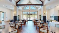 This expansive single-floor community clubhouse offers a generous footprint thoughtfully designed for gathering, dining, and relaxation. The architectural centerpiece is a dramatic vaulted ceiling adorned with bold, dark-stained wood beams that rise to impressive heights, paired with large clerestory windows that flood the interior with natural light and create an airy, open atmosphere.

The dining area features a herringbone-patterned hardwood floor and is lined with neatly arranged tables and cushioned chairs, all bathed in natural light streaming through floor-to-ceiling windows and glass doors that open to an outdoor courtyard. The living area offers comfortable lounge seating and connects seamlessly to a stylish bar nook, highlighted by a striking marble-topped island with a navy blue base, warm wood cabinetry, botanical-print wallpaper, and layered gold pendant lighting.

A welcoming entry lobby showcases soaring ceilings, a decorative laser-cut screen partition, tile-clad accent walls, and a central console table with curated décor. Additional amenity spaces include a game room with teal-painted walls, wall-mounted televisions, and a dedicated card table. Throughout the space, floor-to-ceiling glazing provides seamless views of the surrounding outdoor amenity areas, reinforcing a bright, resort-inspired ambiance.