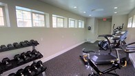 This single-floor fitness room offers a practical and well-appointed space for exercise. The room features light-colored walls that contribute to a clean, open atmosphere, complemented by a row of clerestory windows positioned near the ceiling along one wall, allowing natural light to filter in while maintaining privacy. Recessed ceiling lighting ensures consistent illumination throughout the space. The flooring consists of durable rubber or epoxy-speckled material, well-suited for heavy equipment and high-impact activity. A dumbbell rack stocked with a range of free weights lines one side of the room, while cardio equipment — including a rowing machine and a stationary bike — occupies the central and far areas of the space. An emergency exit sign is visible near the ceiling, and a mounted device, likely a TV or speaker, is affixed to one wall. The overall design is functional and straightforward, prioritizing usability and safety with clean lines, minimal décor, and ample floor space for movement and exercise routines.