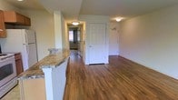 This single-floor space is designed with a functional and welcoming layout, centered around a open living area that connects seamlessly to other parts of the home. The flooring throughout is warm-toned hardwood, lending a cohesive and polished appearance to the interior.

A standout feature is the kitchen breakfast bar, topped with granite countertop in earthy tones and supported by a clean white pedestal base. This bar serves as a natural divider between the kitchen and the main living area, making it ideal for casual dining or entertaining.

A central hallway extends from the main living space, leading to additional rooms. Flush-mounted ceiling lights illuminate the corridor and main area, maintaining a bright and well-lit environment throughout. Multiple paneled white doors line the hallway and open areas, providing access to separate rooms. Natural light is visible at the far end of the hallway, suggesting a window that draws daylight into the interior.

The walls are finished in a soft, neutral tone, complementing the warm wood floors and contributing to the overall clean, understated aesthetic of the space.