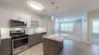This single-floor residence showcases a thoughtfully designed open-concept layout that connects the kitchen and living areas into one cohesive space. The kitchen is a standout feature, equipped with stainless steel appliances including a gas range, microwave, dishwasher, and refrigerator. Two-tone cabinetry pairs white upper cabinets with dark wood-grain lower cabinets, complemented by a white subway tile backsplash and light-toned countertops. A granite-topped center island with pendant lighting above serves as a natural focal point, offering additional prep space and casual seating. Light wood-style flooring runs throughout the kitchen and main living area, contributing to a clean, contemporary aesthetic. The living space receives natural light through a large window fitted with horizontal blinds. The bedroom is carpeted in neutral tones and includes a walk-in closet outfitted with wire shelving for organized storage. The bathroom features light tile flooring, a tub-shower combination, brushed nickel fixtures, a grab bar for added accessibility, and a vanity with a granite countertop. Throughout the unit, a neutral color palette of soft grays and whites reinforces a calm, modern ambiance.