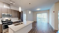 This thoughtfully designed 2-bedroom, 2-bathroom residence offers a seamless open-concept layout that maximizes both functionality and comfort. The kitchen serves as the centerpiece of the living area, featuring rich dark wood cabinetry, a white quartz waterfall island with an undermount sink, a white subway tile backsplash, and a full suite of stainless steel appliances including a gas range, microwave, and French door refrigerator. Elegant pendant lighting illuminates the island, complementing the track lighting overhead. Hardwood-style flooring flows throughout the main living areas, leading to a spacious living room with direct access to an outdoor area through sliding glass doors fitted with vertical blinds. The bedrooms are carpeted for comfort, with the primary bedroom offering multiple windows with outdoor tree views and a ceiling fan. Each bedroom is supported by its own closet with wire shelving systems. Both bathrooms are finished with quartz countertops, wood-toned vanities, large mirrors, and tile flooring. One bathroom features a tub-shower combination with floor-to-ceiling tile surround, while the other offers a dual-sink vanity with direct access to the primary bedroom. The overall aesthetic is clean, modern, and move-in ready.