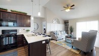 This two-floor home is thoughtfully designed to maximize both style and functionality. The main living area showcases an open-concept layout where the kitchen flows effortlessly into the living room, creating a welcoming and connected space ideal for everyday living and entertaining. The kitchen is appointed with rich dark cabinetry, a light-toned countertop island with bar seating, a double-basin sink, and stylish pendant lighting that adds a warm, modern touch. The living room benefits from soaring vaulted ceilings that lend an impressive sense of volume and openness, while a ceiling fan ensures year-round comfort. Natural light fills the space, highlighting the wood-look flooring that runs throughout the main level, providing warmth and continuity. Crisp white walls and neutral tones create a bright, airy ambiance that serves as a versatile backdrop for any décor style. Decorative wall accents, including a sunburst mirror detail positioned near the ceiling, add character and visual interest to the living area. The two-floor layout offers a clear separation between communal and private spaces, making the home as practical as it is inviting.
