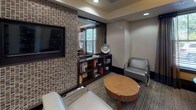 This thoughtfully designed single-floor space blends a library lounge with a comfortable social area, creating a refined yet welcoming environment. The corridor-style library is lined with dark-framed, open built-in shelving units stocked with books and decorative objects, punctuated by structural columns that add architectural rhythm. Overhead, distinctive wood-veneer drum pendant lights cast warm, ambient illumination that complements the recessed ceiling lights throughout.

A textured mosaic tile accent wall in earthy tones of brown and beige serves as a prominent design feature, housing a wall-mounted flat-screen television and framing a large mirror. Beneath it, a dark wood console with open shelving provides additional storage and display space. The seating area includes sleek chrome-framed upholstered chairs and a rounded wood coffee table, creating an intimate retreat.

Adjacent to this area, a casual dining nook features a round table paired with tufted and patterned chairs alongside a built-in upholstered banquette. Large windows with tailored drapery allow natural light to filter in, offering views of the surrounding outdoor greenery. Neutral-toned carpeting unifies the space, while dark wood trim and baseboards provide polished contrast throughout.