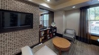 This thoughtfully designed single-floor space blends a library lounge with a comfortable social area, creating a refined yet welcoming environment. The corridor-style library is lined with dark-framed, open built-in shelving units stocked with books and decorative objects, punctuated by structural columns that add architectural rhythm. Overhead, distinctive wood-veneer drum pendant lights cast warm, ambient illumination that complements the recessed ceiling lights throughout.

A textured mosaic tile accent wall in earthy tones of brown and beige serves as a prominent design feature, housing a wall-mounted flat-screen television and framing a large mirror. Beneath it, a dark wood console with open shelving provides additional storage and display space. The seating area includes sleek chrome-framed upholstered chairs and a rounded wood coffee table, creating an intimate retreat.

Adjacent to this area, a casual dining nook features a round table paired with tufted and patterned chairs alongside a built-in upholstered banquette. Large windows with tailored drapery allow natural light to filter in, offering views of the surrounding outdoor greenery. Neutral-toned carpeting unifies the space, while dark wood trim and baseboards provide polished contrast throughout.