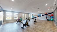 This single-floor fitness studio is purpose-built for cycling and group exercise, offering a spacious, open layout with light wood-look flooring that provides a clean, modern aesthetic throughout. Multiple stationary spin bikes are arranged to allow ample room for movement, making the space well-suited for both individual workouts and group sessions.

One wall features a bold graphic design in navy blue and orange stripes, serving as a dynamic focal point anchored by a large wall-mounted display screen that offers on-demand workout content. Stability balls are stored nearby, adding versatility for cross-training. Additional fitness accessories, including weighted balls in various sizes, are arranged along another wall for easy access.

Natural light enters through a row of large French-style doors and windows along one side of the studio, creating a bright and energizing atmosphere. Recessed ceiling lighting supplements the natural light, ensuring consistent illumination throughout. A glass-panel wall and double-door entry connect the studio to an adjacent space, maintaining an open, connected feel.

The opposite wall is finished in a deep navy blue and houses two restroom facilities, offering practical convenience for users. The overall design balances energetic visual elements with functional amenities, making this a well-equipped and inviting fitness environment.
