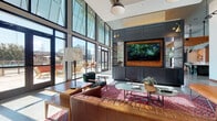 This single-story clubhouse is a thoughtfully designed communal space that blends modern architecture with eclectic, Texas-inspired interiors. Soaring ceilings and expansive floor-to-ceiling glass walls define the main lounge area, flooding the space with natural light while offering views of the surrounding outdoor patio. A tufted leather sofa, patterned area rugs, and a large wall-mounted television framed in warm wood anchor this inviting gathering zone, complemented by dark paneled walls and curated accent lighting.

A separate seating area features a rustic-industrial aesthetic, with a distinctive green sliding barn door, open shelving displaying decorative objects, and a mix of mid-century and woven accent chairs beneath double-height ceilings. A framed Texas map adds a regional touch to this lounge corner.

The dining area accommodates multiple seating arrangements on patterned cement tile flooring, surrounded by natural light from large windows and multiple exterior doors leading to an outdoor terrace. Industrial pendant lights hang above communal tables suited for casual gatherings.

The kitchen is a standout feature, showcasing a large quartz-topped island with teal subway tile detailing, a bold patterned tile backsplash, exposed brick, warm wood ceiling beams, and copper-toned bar stools — creating a vibrant, functional space for cooking and entertaining.