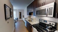 This single-floor space showcases a well-appointed galley-style kitchen as its centerpiece, designed with rich dark espresso cabinetry that extends from floor to ceiling, maximizing storage capacity. The countertops are finished in light granite, offering a striking contrast against the dark wood tones. Stainless steel appliances, including a range, built-in microwave, and dishwasher, lend a sleek, modern aesthetic throughout the kitchen. A subway tile backsplash in a classic white adds texture and a clean visual element to the cooking area. The kitchen flows seamlessly toward a living area, which benefits from abundant natural light streaming through a window and glass door that appear to lead to an outdoor balcony. The flooring throughout is a warm-toned hardwood, unifying the kitchen and living spaces cohesively. Crown molding along the ceiling adds an architectural detail that elevates the overall finish of the space. A tall mirror near the transition between rooms enhances the sense of openness, reflecting light and adding visual depth to the interior.