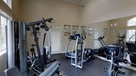 This fitness room offers a practical and well-organized workout environment within a single-level space. The room features classic architectural detailing, including crown molding along the upper perimeter and a chair rail that divides the warm beige walls, lending the space a refined character uncommon in typical gym settings. Large mirrors line one wall, visually expanding the room and allowing users to monitor their form during exercise. The flooring consists of durable dark rubber matting, suitable for heavy equipment and high-impact activities. Equipment on offer includes a multi-function cable weight machine, at least two elliptical cross-trainers, and a treadmill, providing options for both strength training and cardiovascular workouts. Informational exercise posters are displayed along the walls, serving as handy visual references. Two white paneled doors provide access points, and the neutral lighting creates a clean, functional atmosphere throughout. The overall ambiance balances traditional architectural charm with a purpose-built fitness environment.