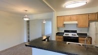 This single-floor space offers a practical and well-connected layout designed for comfortable everyday living. The kitchen serves as a central highlight, featuring dark granite countertops that wrap around a peninsula breakfast bar, warm honey-toned wood cabinetry, and a gas range with overhead ventilation hood. Recessed and flush-mount ceiling lighting keeps the kitchen bright, while two elegant pendant lights hang above the peninsula, adding a refined touch to the space.

The open-concept design allows the kitchen to flow naturally into the adjacent living area, which is connected through a wide passageway. Built-in shelving units flank the transition between spaces, offering generous storage and display options. Sliding closet doors and thoughtful built-ins contribute to the space's organizational functionality.

The living area extends the sense of openness, with neutral-toned carpet flooring that creates a warm contrast to the kitchen's hard surfaces. The overall color palette throughout the space is light and neutral, with soft beige walls that reflect available light and lend an airy, inviting ambiance. The layout is cohesive and efficient, making effective use of every area across the single floor to deliver a functional and comfortable living environment.