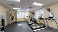 This fitness center is designed to support a comprehensive workout experience within a compact, single-level layout. The room features a drop-panel ceiling with recessed fluorescent lighting, warm beige walls, and durable dark rubber flooring throughout, creating a clean and functional environment. Natural light enters through multiple windows fitted with white horizontal blinds, enhancing the bright atmosphere alongside the overhead lighting.

The cardio section is well-appointed with multiple True-brand treadmills and elliptical trainers, along with a stationary cycle, providing ample options for aerobic exercise. A wall-mounted flat-screen television is positioned for viewing during workouts. A wall-mounted fan offers additional air circulation.

The strength training area includes a multi-functional cable machine and a dedicated weight bench paired with a rack of dumbbells in graduated sizes. A selectorized leg press/extension machine provides additional resistance training options. For functional fitness, the space offers a medicine ball rack stocked with various weighted balls, stability balls, and a water cooler station for hydration. Two entry doors provide convenient access. The overall layout is organized and spacious, allowing users to move freely between equipment zones.
