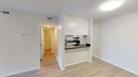 This single-story unit is designed with a clean, modern aesthetic throughout, combining practical functionality with bright, open interiors. The living area serves as the centerpiece of the space, featuring light wood-look laminate flooring, a ceiling fan with light fixture, and a sliding glass door dressed with vertical blinds that floods the room with natural light and provides outdoor access. A thermostat and dedicated HVAC venting ensure year-round climate comfort.

The kitchen is thoughtfully positioned behind a pass-through opening, offering visual connectivity to the main living area. It features dark countertops, white cabinetry, a stainless steel sink, a refrigerator, and a range, along with a subtle subway tile backsplash that adds a contemporary touch. Light wood-look flooring continues seamlessly from the living space into the kitchen.

The bedroom is carpeted for comfort and benefits from its own ceiling fan and a window that allows natural light to filter in. A hallway connects the living spaces to the bedroom and bathroom, maintaining an efficient and logical layout.

The bathroom is well-appointed with a wide vanity featuring white cabinetry, a large mirror with overhead lighting, and a tub-shower combination set beneath a distinctive arched surround with a curtain rod. Wood-look flooring ties the bathroom's design to the rest of the unit, contributing to a cohesive and polished overall ambiance.