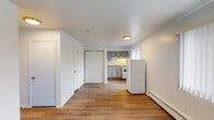 This single-floor unit offers a clean, functional layout designed for efficient everyday living. The main living area is finished with warm wood-tone laminate flooring that extends throughout the space, complemented by crisp white walls and flush-mount ceiling lighting. A baseboard heating system runs along the perimeter walls, providing consistent warmth throughout. The open-plan design connects the living area directly to the kitchen, which features gray shaker-style cabinetry with brushed nickel hardware, granite-patterned countertops, a stainless steel sink, a white gas range with hood ventilation, and a full-size white refrigerator. A window above the kitchen area allows natural light to filter in. Storage is addressed with a built-in closet unit accessible from the main living space, along with an additional interior door leading to further rooms. The bedroom is a well-proportioned, bright room with a large window fitted with vertical blinds and a baseboard heater beneath it. The full bathroom includes a tub-shower combination with tan tile surround and a dark curtain, a white vanity with sink, a toilet, a mirrored medicine cabinet, and a lighted vanity fixture above. The overall ambiance is clean, minimal, and move-in ready.