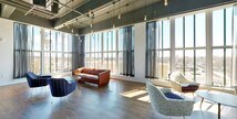This single-floor space blends industrial design with contemporary furnishings to create a versatile, open-plan lounge and event venue. The most defining architectural feature is the wraparound wall of floor-to-ceiling windows, which floods the interior with abundant natural light and offers sweeping elevated views of the surrounding cityscape and skyline. Exposed ductwork, structural beams, and a dark painted ceiling reinforce the industrial aesthetic, while warm hardwood flooring throughout softens the overall tone.

The space is organized into several distinct functional zones. A lounge area features an eclectic mix of seating, including a tufted leather sofa, velvet accent chairs, and a low oval coffee table, all arranged to encourage relaxed conversation. Sheer draped curtains line the windows, adding a refined texture while preserving the views. Adjacent to the lounge, a high-top dining and gathering area provides multiple bistro-style tables paired with sleek bar stools, suitable for casual dining or social events.

A full-service bar anchors another section of the space, finished with a striking teal geometric tile facade, a dark countertop, and lined with retro chrome bar stools. Mounted televisions and additional seating complete this area. Glass-panel doors open to an outdoor terrace, extending the venue's entertaining capacity and further connecting the interior to the panoramic views beyond.