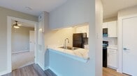 This efficient and thoughtfully laid-out one-bedroom, one-bathroom residence makes excellent use of its compact footprint. The living area features durable hardwood-style flooring in a cool gray tone, warm neutral-painted walls, and a large window with slatted blinds that invites generous natural light. A private entry door adds a welcoming touch to the space.

The kitchen is designed with a pass-through counter featuring a striking marble-style countertop and a stainless steel double sink, providing both a functional workspace and an open connection to the main living area. White shaker-style cabinetry lines the kitchen walls, complemented by black appliances including a refrigerator and range, creating a clean, modern contrast.

The bedroom offers plush wall-to-wall carpeting for added comfort underfoot, a ceiling fan with integrated lighting for year-round comfort, and a well-sized window that fills the room with natural light. A dedicated closet provides convenient storage. The hallway connecting the rooms features matching hardwood-style flooring that maintains a cohesive flow throughout the home.

The bathroom is finished with a full soaking tub and shower combination, surrounded by large-format, light-toned tile in a soft linear pattern, lending the space a clean and spa-like aesthetic. Overall, the residence presents a bright, neutral palette that is both practical and inviting.