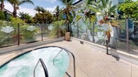 This outdoor amenity space offers a well-appointed resort-style experience centered around a generously sized rectangular swimming pool with clear, blue water. The pool deck is lined with lounge chairs and shaded by blue market umbrellas, providing ample space for relaxation in the sun. The surrounding buildings feature a modern design with bold blue exterior cladding and open-air corridors, creating a vibrant and contemporary backdrop for the outdoor area.

Tall palm trees and tropical plantings frame the space, lending a warm, Southern California ambiance. A dedicated spa and hot tub area is tucked adjacent to the main pool, enclosed by a glass panel fence with decorative frosted designs for privacy and safety. This intimate space is surrounded by lush tropical foliage, including banana leaf plants and palms, creating a secluded, garden-like retreat.

The pool deck is finished with light-toned concrete surfacing, and the entire amenity area is enclosed by a perimeter fence, ensuring a secure environment. Overall, the space combines modern architectural elements with tropical landscaping to create an inviting and functional outdoor setting ideal for recreation and leisure.