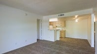 This single-floor unit presents an open-concept layout that connects the living area to the kitchen, creating a cohesive and functional living space. The living area is finished with wall-to-wall carpeting in a neutral tan tone, offering a comfortable and understated foundation that complements the white walls throughout.

The kitchen is positioned centrally and features a peninsula-style breakfast bar with an overhanging countertop, providing casual seating and serving as a natural divider between the kitchen and living space. Light wood cabinetry lines the upper and lower sections of the kitchen, offering ample storage. A white range is integrated beneath the upper cabinets, accompanied by an under-cabinet range hood. Overhead fluorescent lighting illuminates the workspace effectively.

A ceiling fan is visible in the adjoining room beyond the kitchen, suggesting an additional living or dining area. A doorway with a bi-fold door, likely leading to a closet, is visible along the living room wall. The textured popcorn ceiling and HVAC ceiling vent are consistent throughout, contributing to the unit's practical and clean aesthetic. Overall, the space maintains a simple, functional character with a neutral palette.