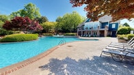 Located at 1701 Dawsonville Hwy in Gainesville, Georgia, this outdoor amenity complex offers a thoughtfully designed resort-style retreat. The centerpiece is a large, freeform swimming pool with striking brick-edged coping and crystal-clear blue water, complemented by a separate spa pool and a decorative fountain feature. An expansive concrete pool deck surrounds the water, lined with rows of lounge chairs ideal for sunbathing and relaxation.

The grounds are meticulously landscaped with a rich variety of mature trees, including vibrant Japanese maples, ornamental shrubs, and manicured hedgerows that provide both color and privacy throughout the seasons. A brick pergola with a slatted overhead canopy anchors a secondary gathering area, accessible via curved brick staircases that add classical architectural character to the outdoor space.

A substantial two-story clubhouse building with blue siding, brick columns, large windows, and an upper-level balcony overlooks the pool area, providing a prominent architectural backdrop to the complex. Additional lounge seating is arranged near the building's ground-floor entrance.

Beyond the pool deck, open lawn areas and winding pathways extend into beautifully maintained gardens, opening to sweeping views of the rolling Georgia hills under wide, open skies — making this outdoor space as scenic as it is functional.