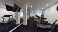 Located at 5855 Carlton Way in Los Angeles, CA, this fitness center offers a well-equipped, single-floor workout facility designed for functionality and comfort. The space features a clean, modern aesthetic with crisp white walls, dark rubber flooring, and recessed lighting throughout, creating a focused and energizing environment.

The cardio zone is well-stocked with multiple treadmills and elliptical trainers from Spirit and other brands, as well as stationary bikes — providing a comprehensive range of aerobic training options. A dedicated strength and free weights area includes a full rack of dumbbells in varying weights, medicine balls, and stability balls, catering to a wide range of fitness routines.

Full-length mirrors line the walls, enhancing the sense of space and allowing users to monitor their form during workouts. A wall-mounted flat-screen TV is positioned for easy viewing from the cardio machines. The facility is naturally lit by windows and supplemented by recessed ceiling lights, maintaining a bright and open atmosphere.

A flat workout bench near the entrance provides a convenient space for stretching or accessory exercises. The room is accessible via a secure entry door and is equipped with emergency exit signage, ensuring safety. The overall design prioritizes practicality while offering a complete fitness experience in the heart of Los Angeles.