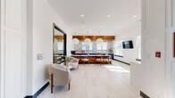 The lobby at 1003 148th St W in Rosemount, Minnesota, offers a well-appointed, single-floor communal space that blends modern design with warm, welcoming details. Upon entry, a striking coffered ceiling accent in teal blue, adorned with an elegant chandelier, sets the tone for the refined interior. Large-format light stone tile flooring extends throughout, creating a clean and cohesive aesthetic.

The open layout seamlessly connects a comfortable lounge area — featuring a curved upholstered chair and a framed oversized mirror — with a fully equipped kitchen workspace. The kitchen is a design standout, showcasing bold cobalt blue cabinetry, a warm wood-paneled island with a crisp white quartz countertop, and a warm wood soffit overhead. A geometric teal mosaic tile backsplash adds a vibrant pop of color, complemented by large pendant light fixtures. Ample natural light pours in through multiple windows, offering views of the surrounding neighborhood.

A dining area with mid-century modern wood chairs sits adjacent to the kitchen, ideal for meetings or communal gatherings. A wall-mounted flat-screen TV and hardwood-floored corridor connect the space to additional areas of the building. The overall ambiance is contemporary and polished, balancing functionality with a welcoming, upscale atmosphere.