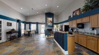 This single-floor community space makes a striking first impression with its soaring vaulted ceilings and bold teal and white color scheme that carries throughout. The entryway welcomes visitors with rich slate tile flooring, decorative columns, and large windows that frame the entrance, flooding the space with natural light.

The open main area flows seamlessly between distinct functional zones. A comfortable lounge area centers around a tile-surround fireplace, accented by teal upholstered chairs, a round wood coffee table, and a large vibrant abstract painting mounted prominently above the mantle. Teal drapery panels frame the windows, adding warmth and visual cohesion.

Adjacent to the lounge, a formal dining area features a dark wood table with cream upholstered chairs, surrounded by floor-to-ceiling windows dressed in matching teal drapes, offering an inviting and private setting.

A service counter with decorative wrought iron detailing defines the leasing or reception area, complemented by nearby workstations. A dedicated kitchenette provides ample cabinetry, granite-style countertops, stainless steel appliances including a full-size refrigerator, a coffee station, microwave, and sink — well-equipped for staff and guests alike.

Throughout the space, carefully chosen artwork, lush indoor plants, and recessed lighting contribute to a polished, welcoming ambiance.