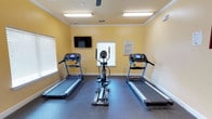 This single-floor fitness room offers a focused workout environment designed for cardiovascular exercise. The space is finished with warm yellow walls accented by white crown molding, creating a bright and energetic atmosphere conducive to exercise. Durable dark rubber flooring covers the workout area, providing both safety and stability for equipment use.

The room is equipped with two treadmills positioned on either side of a central elliptical cross-trainer, offering users a variety of low-impact and higher-intensity cardio options. Each treadmill features an integrated digital display console for tracking workout metrics. A wall-mounted flat-screen television is positioned at an optimal viewing height, allowing users to stay entertained during their sessions.

Natural light filters in through multiple windows fitted with horizontal blinds, while overhead fluorescent lighting ensures the space remains well-lit at all times. A ceiling-mounted fan aids in air circulation, helping maintain a comfortable temperature during workouts. Fitness center rules are posted on the wall, ensuring users are informed of proper conduct and equipment usage guidelines.