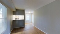 This 1-bedroom, 1-bathroom single-floor unit is thoughtfully designed to maximize functionality within a cozy footprint. The open-concept living area features warm wood-look flooring that runs throughout, creating a cohesive and inviting atmosphere. Neutral gray accent walls complement the light, airy tones of the space, lending a modern yet approachable aesthetic. A kitchen pass-through window connects the cooking area to the living space, promoting an open feel while defining each zone. The kitchen is equipped with a range and overhead exhaust hood, with white cabinetry providing clean contrast against the darker accent wall. The living area benefits from natural light streaming through a window at the far end, brightening the space and creating a welcoming ambiance. A textured ceiling adds subtle visual character throughout. The layout flows efficiently from the entry through the living area, making smart use of every square foot. Clean white baseboards and simple, understated finishes give the unit a well-maintained and move-in-ready appearance, ideal for those seeking a straightforward and comfortable living space.