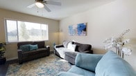 This single-floor, one-bedroom, one-bathroom space offers a comfortable and well-appointed layout designed for everyday living. The living room serves as the centerpiece of the unit, featuring a ceiling fan with a light fixture that provides both air circulation and ambient illumination. Neutral, warm-toned walls create a clean backdrop that complements the space's relaxed aesthetic.

Natural light filters in through a window fitted with horizontal blinds, brightening the room and highlighting the patterned area rug in soft blues and earth tones that anchors the seating arrangement. The living area comfortably accommodates multiple seating pieces, including a striped sofa with decorative pillows and an upholstered armchair, contributing to a cozy, inviting atmosphere.

A floral canvas print in shades of blue and blush adds a decorative focal point on the wall, tying together the room's cool, calming color scheme. A floor lamp provides supplemental lighting, enhancing the warmth of the space during evening hours. Overall, the unit presents a tidy, move-in-ready environment with a light, airy feel throughout.