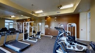 This fitness center offers a comprehensive workout environment designed to accommodate a range of exercise needs. The space is outfitted with multiple strength training cable machines and a stationary exercise bike, providing options for both resistance and cardiovascular training. The flooring consists of durable, patterned carpet suited for a gym setting, while the walls are finished in warm earth tones, giving the room an inviting atmosphere. Full-length mirrors line one wall, creating a sense of expanded space and allowing users to monitor their form during workouts. Overhead, ceiling fans work in tandem with recessed lighting and fluorescent fixtures to maintain a comfortable and well-lit environment. Ventilation is supported by visible HVAC vents integrated into the ceiling. The layout is thoughtfully arranged to allow clear movement between equipment. The combination of functional design, warm finishes, and practical amenities makes this fitness center a well-rounded facility suitable for daily use.