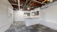 This one-bedroom, one-bathroom unit showcases a compelling blend of industrial character and modern updates throughout its efficiently designed single-floor layout. The open-ceiling design reveals original wood beams, exposed ductwork, and piping overhead, lending an authentic loft-like aesthetic that defines the overall ambiance of the space.

The main living area is anchored by a peninsula-style kitchen featuring dark gray cabinetry with brushed nickel hardware, white quartz countertops, white appliances including a range, microwave, and refrigerator, and a built-in sink. A washer unit is also integrated near the kitchen peninsula. Patterned carpet tiles in muted gray tones cover the floors throughout most of the unit, while the kitchen transitions to light wood-look flooring. Two windows bring in natural light to the living area, complemented by a ceiling fan overhead.

Two hallways connect the rooms, with one showcasing a striking painted white brick wall. The bedroom features exposed red brick walls, a ceiling fan, a window with natural light, and a cast iron radiator. The bathroom is a standout space, with original red brick walls, large-format tile flooring, a walk-in tiled shower with a built-in bench, and a small window for ventilation.