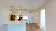 This single-floor space showcases a clean, contemporary design with an emphasis on functionality and modern aesthetics. The open-concept kitchen serves as a central highlight, featuring crisp white shaker-style cabinetry that extends to the upper walls, providing generous storage. Stainless steel appliances, including a refrigerator and a gas range with an overhead microwave, offer a polished, professional-grade cooking environment. A sleek white quartz countertop island anchors the kitchen, providing additional workspace and serving as a natural gathering point. Elegant pendant lighting hangs above the island, adding a refined touch to the space. The kitchen flows seamlessly into the adjacent living area, creating an open and connected layout ideal for both daily living and entertaining. Warm hardwood-style flooring runs consistently throughout, lending a cohesive and inviting feel to the interior. The entryway is clearly defined with a solid door and keypad entry, complemented by a utility panel nearby. Recessed lighting and ceiling-integrated HVAC vents ensure comfort and efficient climate control. The light-toned walls and bright finishes throughout contribute to an airy, well-lit ambiance that feels both welcoming and polished.