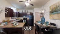 This single-floor space showcases an open-concept layout that seamlessly connects the kitchen and dining area, creating a cohesive and functional living environment. The kitchen is a standout feature, outfitted with rich dark wood cabinetry that extends to the upper walls, providing generous storage. Granite-style countertops add a refined aesthetic, complementing the dark tones of the cabinetry and black appliances, which include a refrigerator, range, and microwave. A kitchen island extends the counter space and serves as a casual gathering point. Wood-look flooring runs throughout, adding warmth and visual continuity across both the kitchen and dining areas. The dining space comfortably accommodates a table and chairs, making it well-suited for everyday meals or entertaining. A ceiling fan with an integrated light fixture is centrally positioned overhead, ensuring both air circulation and ambient lighting. The neutral wall tones provide a clean backdrop that allows the darker furnishings and finishes to take center stage. The overall ambiance is warm and inviting, balancing practical design with polished finishes.