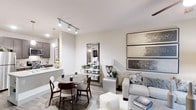 This well-appointed single-floor residence offers a seamlessly connected open-concept layout that integrates the living, dining, and kitchen areas into one cohesive and functional space. The interior is defined by a neutral color palette of soft grays and whites, creating a calm and contemporary ambiance throughout.

The kitchen features dark cabinetry paired with light stone countertops and is equipped with modern appliances. Pendant lighting above the counter adds a refined touch, while the open pass-through design keeps the kitchen visually connected to the rest of the living area.

The dining area comfortably accommodates a round table with upholstered chairs, enhanced by track lighting overhead that provides both practicality and style. Adjacent to it, the living area is generously proportioned, with plush seating, layered textures, and carefully curated décor including a large decorative mirror and framed wall art that lend the space a polished, elegant character.

Wood-look flooring runs throughout, adding warmth and continuity to the design. The layout flows effortlessly, making the space feel open and inviting. Overall, this residence combines modern aesthetics with practical living, offering a comfortable and visually cohesive environment.