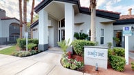 Sunstone's clubhouse makes an immediate impression with its grand exterior entrance, framed by lush tropical landscaping, vibrant flower beds, and towering palm trees. A covered portico leads into a beautifully designed rotunda lobby that serves as the heart of the space. The circular room draws the eye upward to soaring ceilings lined with a ring of clerestory windows, flooding the interior with abundant natural light. Dramatic woven pendant light fixtures descend from the domed ceiling, adding warmth and architectural interest overhead. Large-format hexagonal floor tiles echo the room's circular geometry and lend a clean, modern aesthetic. A curved mosaic-tiled fireplace with a metallic honeycomb pattern serves as a striking focal point on one wall, while a sleek central pedestal table anchored by a vivid teal area rug defines the gathering space. Curved leather seating and colorfully patterned accent chairs are arranged throughout, creating comfortable conversation areas. Artwork adorns the walls, contributing to the vibrant, contemporary ambiance. A glimpse beyond the lobby reveals additional amenity spaces, hinting at the full range of offerings within this thoughtfully designed community clubhouse.