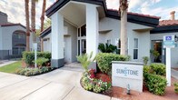 Sunstone's clubhouse makes an immediate impression with its grand exterior entrance, framed by lush tropical landscaping, vibrant flower beds, and towering palm trees. A covered portico leads into a beautifully designed rotunda lobby that serves as the heart of the space. The circular room draws the eye upward to soaring ceilings lined with a ring of clerestory windows, flooding the interior with abundant natural light. Dramatic woven pendant light fixtures descend from the domed ceiling, adding warmth and architectural interest overhead. Large-format hexagonal floor tiles echo the room's circular geometry and lend a clean, modern aesthetic. A curved mosaic-tiled fireplace with a metallic honeycomb pattern serves as a striking focal point on one wall, while a sleek central pedestal table anchored by a vivid teal area rug defines the gathering space. Curved leather seating and colorfully patterned accent chairs are arranged throughout, creating comfortable conversation areas. Artwork adorns the walls, contributing to the vibrant, contemporary ambiance. A glimpse beyond the lobby reveals additional amenity spaces, hinting at the full range of offerings within this thoughtfully designed community clubhouse.
