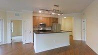 This single-floor residence offers a thoughtfully designed open-concept layout that seamlessly connects the kitchen and living areas. The kitchen serves as the heart of the space, showcasing warm honey-toned wood cabinetry paired with dark granite countertops that provide a striking contrast. A large central island with a white base offers additional workspace and casual seating, making it ideal for both cooking and entertaining. Stainless steel appliances, including a refrigerator, microwave, and range, lend the kitchen a modern, polished aesthetic. Above the island, a decorative pendant light fixture with an integrated pot rack adds a functional yet stylish focal point. A tile backsplash behind the range complements the overall design. Rich hardwood flooring flows throughout the space, adding warmth and continuity to the open floor plan. Crown molding along the ceiling adds an elegant architectural detail. Recessed and track lighting ensure the space is well-lit throughout. A hallway visible from the kitchen leads to additional rooms, hinting at a practical and well-connected floor plan. The overall ambiance is clean, modern, and inviting.
