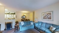 This single-floor space offers a comfortable and cohesive layout, with the living area serving as the central hub of the home. The living room is furnished with plush blue sofas arranged to encourage conversation and relaxation, set atop wall-to-wall carpeting that adds warmth underfoot. Neutral-toned walls keep the space feeling bright and airy, complemented by soft ambient lighting from both a table lamp and a decorative chandelier visible in the adjoining dining area.

The open-concept flow connects the living room seamlessly to the dining space, which features a chandelier overhead and an ornate gold-framed mirror as a focal decorative element, lending a touch of classic elegance. Floral arrangements add a natural, welcoming accent to the dining area.

A glimpse into the kitchen reveals white cabinetry and appliances, suggesting a functional and clean culinary space. Framed wall art in the living room adds a finishing touch, contributing to the overall polished and homey ambiance. The single-floor layout ensures easy navigation throughout, making the space practical and accessible.