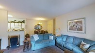 This single-floor space offers a comfortable and cohesive layout, with the living area serving as the central hub of the home. The living room is furnished with plush blue sofas arranged to encourage conversation and relaxation, set atop wall-to-wall carpeting that adds warmth underfoot. Neutral-toned walls keep the space feeling bright and airy, complemented by soft ambient lighting from both a table lamp and a decorative chandelier visible in the adjoining dining area.

The open-concept flow connects the living room seamlessly to the dining space, which features a chandelier overhead and an ornate gold-framed mirror as a focal decorative element, lending a touch of classic elegance. Floral arrangements add a natural, welcoming accent to the dining area.

A glimpse into the kitchen reveals white cabinetry and appliances, suggesting a functional and clean culinary space. Framed wall art in the living room adds a finishing touch, contributing to the overall polished and homey ambiance. The single-floor layout ensures easy navigation throughout, making the space practical and accessible.