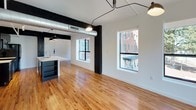 This 2-bedroom, 2-bathroom unit blends industrial character with modern finishes across a well-appointed single-floor layout. Warm natural hardwood floors run throughout, complementing crisp white walls and striking exposed black structural columns and beams that serve as architectural focal points in every room. The open-concept living and dining area is generously proportioned, featuring exposed silver ductwork overhead, pendant and track lighting, and large windows that invite abundant natural light and offer views of the surrounding streetscape and greenery. The kitchen is outfitted with bold dark cabinetry, white countertops, black appliances, and a central island with a sink — a practical and stylish space for cooking and entertaining. Both bedrooms are bright and airy, each equipped with a ceiling fan and multiple windows framing outdoor views. The two bathrooms offer distinct designs: one features classic white subway tile, a soaking tub, and gold-tone fixtures, while the other showcases a glass-enclosed shower, dark vanity cabinetry, a round mirror, and patterned penny tile flooring with black accents. The entryway provides a clean, welcoming arrival point with built-in storage nearby. Throughout, the unit delivers a cohesive industrial-modern aesthetic defined by contrast, warmth, and thoughtful detail.